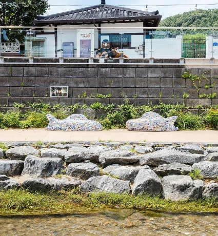 제민천 산책로에 놓인 자갈 모자이크 동물 조형물 두 점. 뒤편 석축 위로 작은 건물이 자리하고 앞쪽 얕은 물가에는 큰 디딤돌이 놓여 있다.