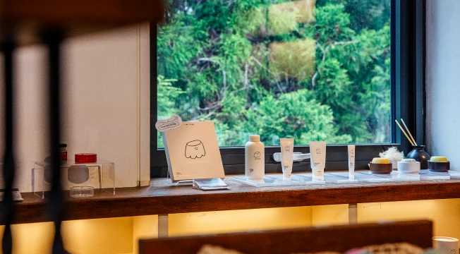 창가 선반에 화장품과 생활용품이 일렬로 진열되고 큰 창 너머 초록 숲이 보이는 모습