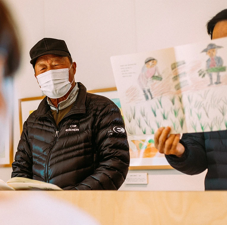 모자와 마스크를 쓴 어르신이 눈을 감고 앉아 있고, 옆에서 손그림 그림책을 펼쳐 보여주는 모습