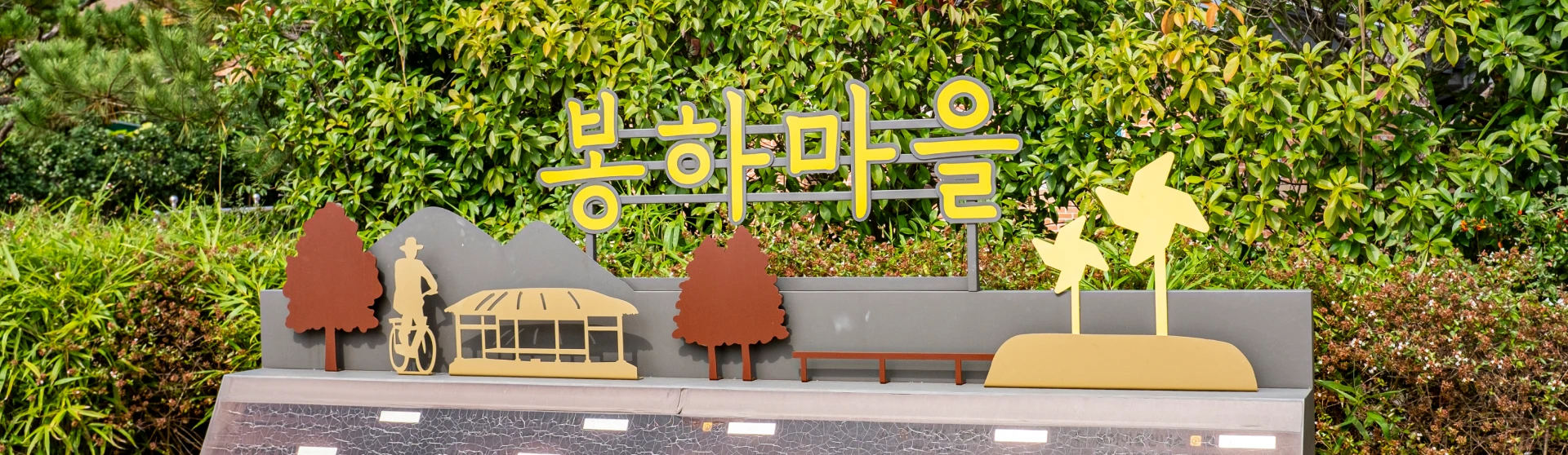 초록 수풀 앞에 노란 봉하마을 글자와 자전거 탄 인물, 나무 모양 조형물이 놓인 마을 입구 표지