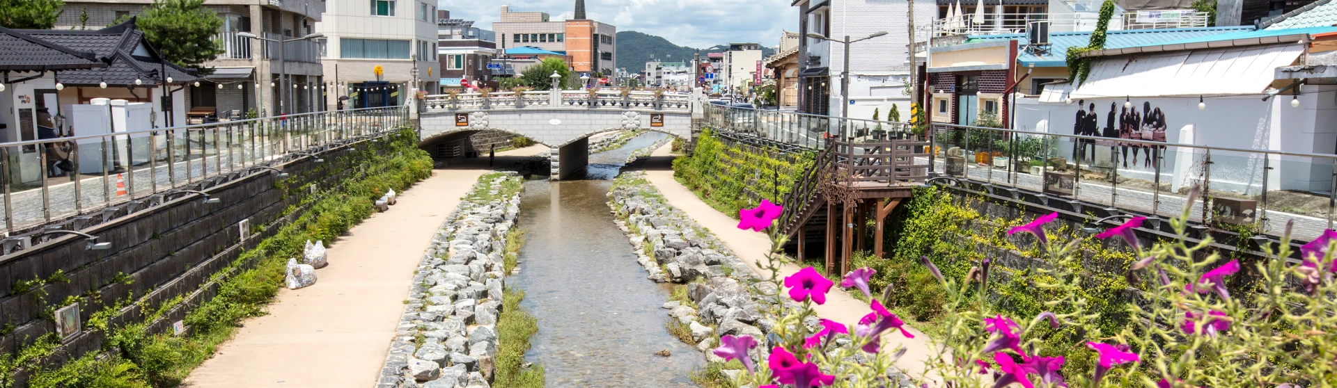 공주 원도심을 가로지르는 제민천 전경. 돌로 만든 아치형 다리 아래로 맑은 물이 흐르고, 하천 양옆 산책로와 옛 건물들이 늘어서 있으며 오른쪽 화단에는 분홍빛 페튜니아가 활짝 피어 있다.