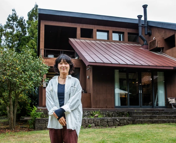 붉은 지붕 목조 건물을 배경으로 잔디밭에 서서 미소 짓는 윤자연 책임 매니저의 전신 모습