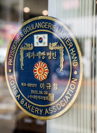 유리문에 붙은 대한제과협회 제과제빵 명인 인증 동판, 명인 이름과 날짜가 새겨져 있다