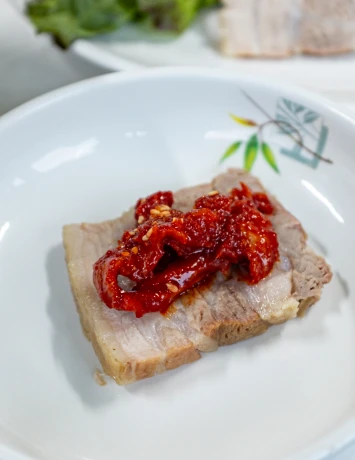 흰 접시에 올린 도톰한 수육 한 점 위에 다진 붉은 양념을 얹은 보쌈 요리 클로즈업