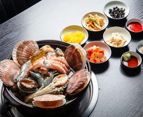 검은 뚝배기에 문어와 가리비, 전복, 새우를 올린 해신탕과 함께 곁들임 반찬 여러 종지가 차려진 한 상