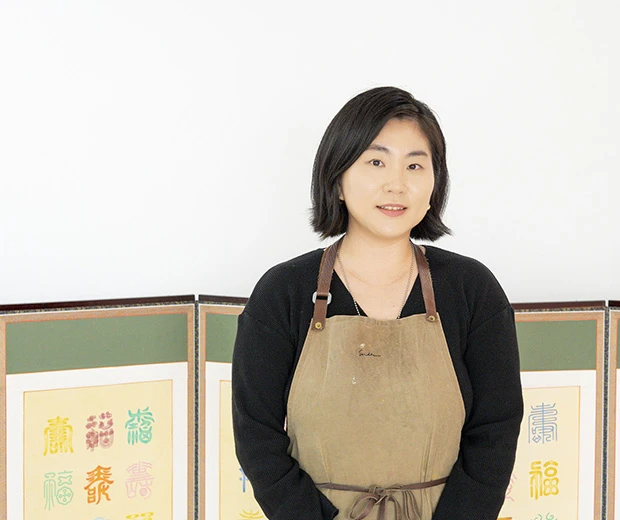 수복 글자 병풍 앞에서 앞치마를 두르고 잔잔히 미소 짓고 있는 루씨손 작가의 모습