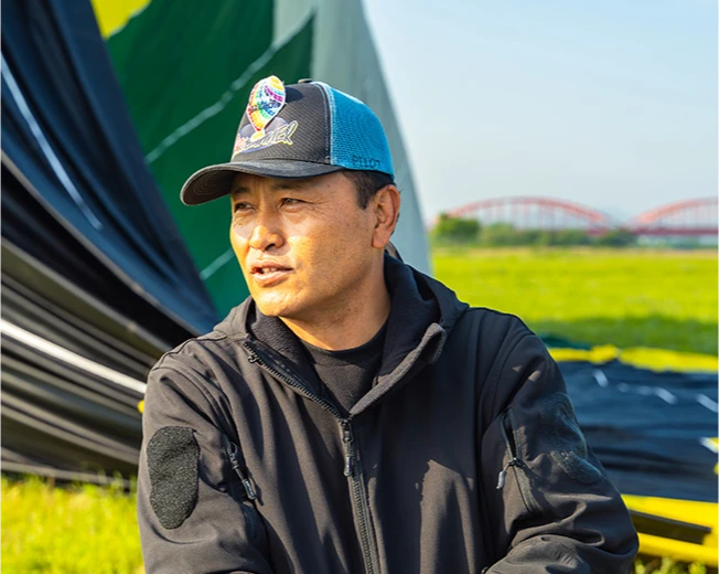 강변에 눕힌 열기구를 배경으로 모자를 쓴 파일럿이 이야기하는 옆모습