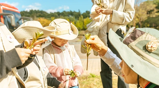 모자를 쓴 아이들과 어른들이 잎이 달린 나뭇가지를 손에 들고 함께 관찰하는 모습