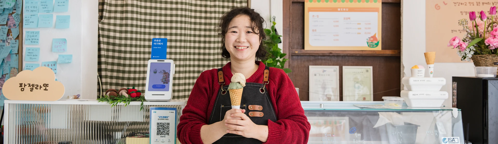 빨간 스웨터에 앞치마를 두른 팜젤라또 대표가 젤라또 콘을 두 손으로 들고 카운터 앞에서 환하게 웃는 모습
