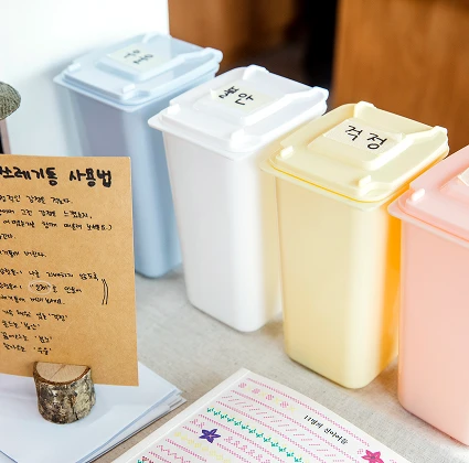 파스텔 색 분리수거함이 나란히 놓여 있고, 옆에는 손글씨로 '쓰레기통 사용법'을 적은 크라프트 안내판과 스티커가 함께 보인다.