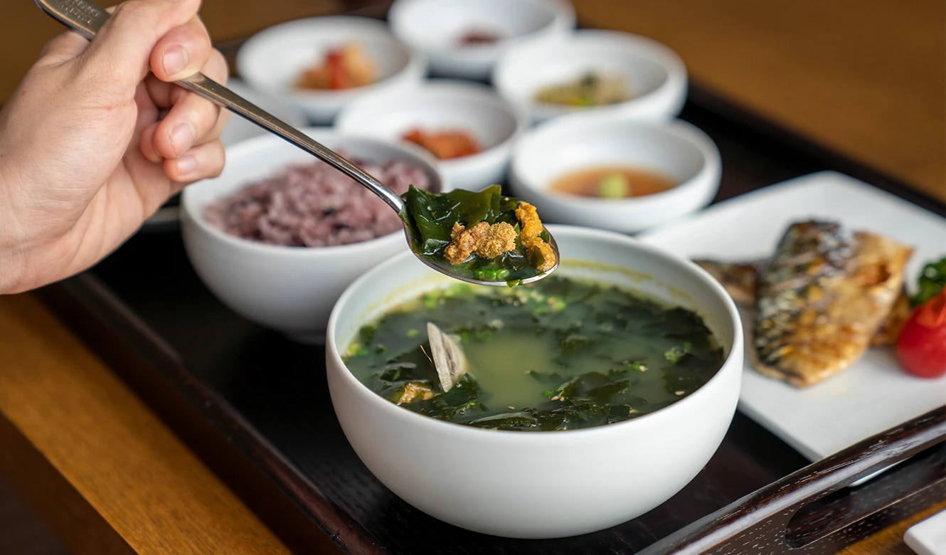 숟가락으로 성게 미역국을 뜨는 손, 흑미밥과 고등어구이, 여러 밑반찬이 정갈하게 차려져 있다