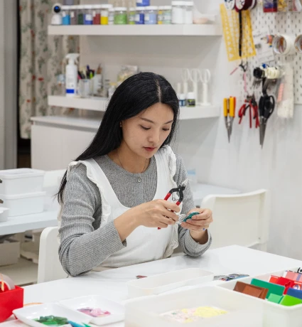 공방 작업대에 앉아 도구로 유리 공예품을 손질하는 긴 머리 여성의 모습