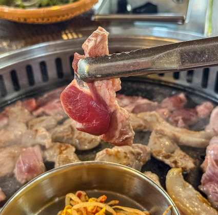 불판에서 구워지는 고기 한 점을 집게로 집어 들어 올린 뒷고기 구이 클로즈업