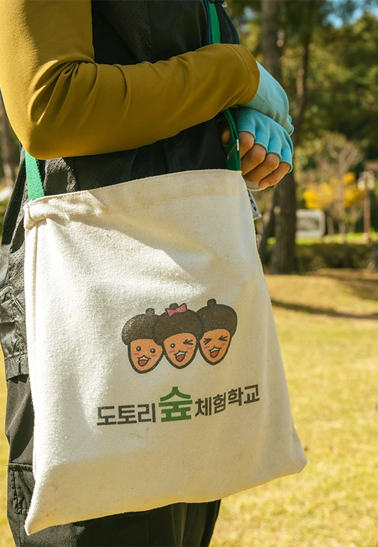 도토리 숲 체험학교 로고가 그려진 에코백을 어깨에 메고 장갑 낀 손으로 가방을 든 모습