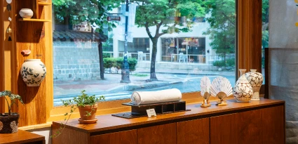 통유리 창가 선반에 도자기와 부채 공예품이 놓이고 창밖으로 하천 풍경이 보이는 공간