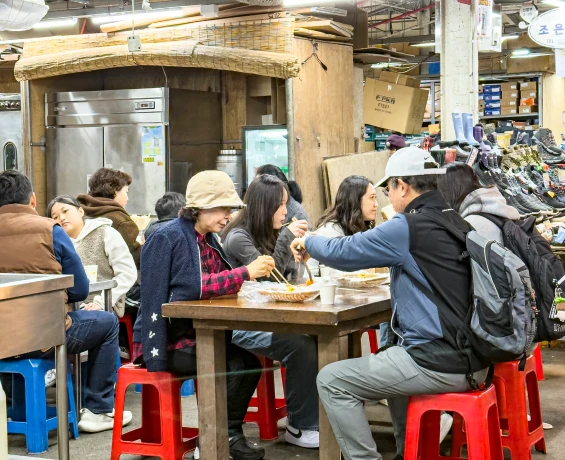 빨간 플라스틱 의자에 둘러앉아 음식을 나누는 시장 안 노포 식당의 활기찬 풍경