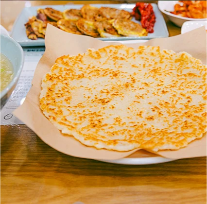 종이 위에 둥글고 노릇하게 부친 큼직한 감자전 한 장과 뒤편의 여러 부침개가 차려진 모습