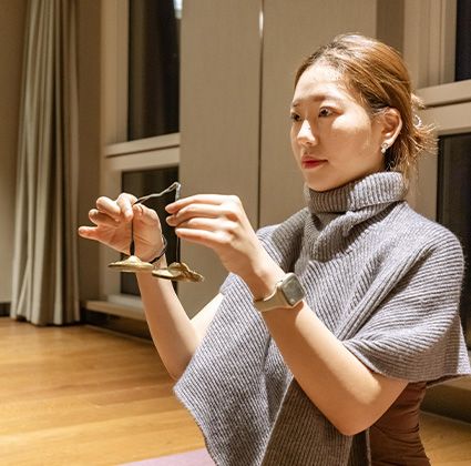 회색 숄을 두른 여성이 손가락으로 작은 명상용 심벌즈를 들고 소리를 울리려 하고 있다