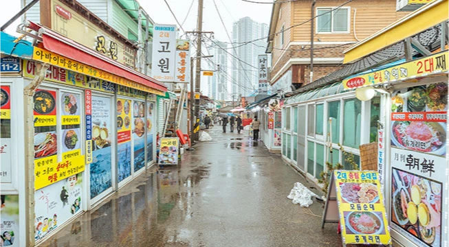 음식 사진 간판이 빼곡한 아바이마을 골목, 비에 젖은 길과 양옆에 늘어선 식당들