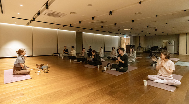 강사와 참가자들이 매트에 마주 앉아 두 손을 모으고 명상에 앞서 인사를 나누고 있다
