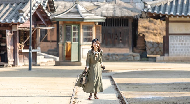 올리브색 개화기 원피스를 입은 여성이 한옥 마당을 거닐며 환하게 웃는 사진 놀이 장면