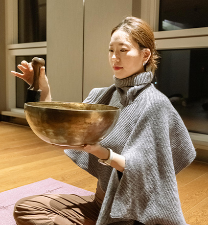 회색 숄을 두른 여성이 큰 싱잉볼을 받쳐 들고 말렛으로 울림을 내려는 순간을 담았다