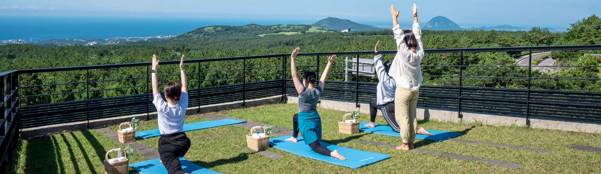 제주 숲과 바다가 내려다보이는 전망대 잔디에서 참가자들이 파란 요가 매트에 서서 두 팔을 위로 뻗는 모습