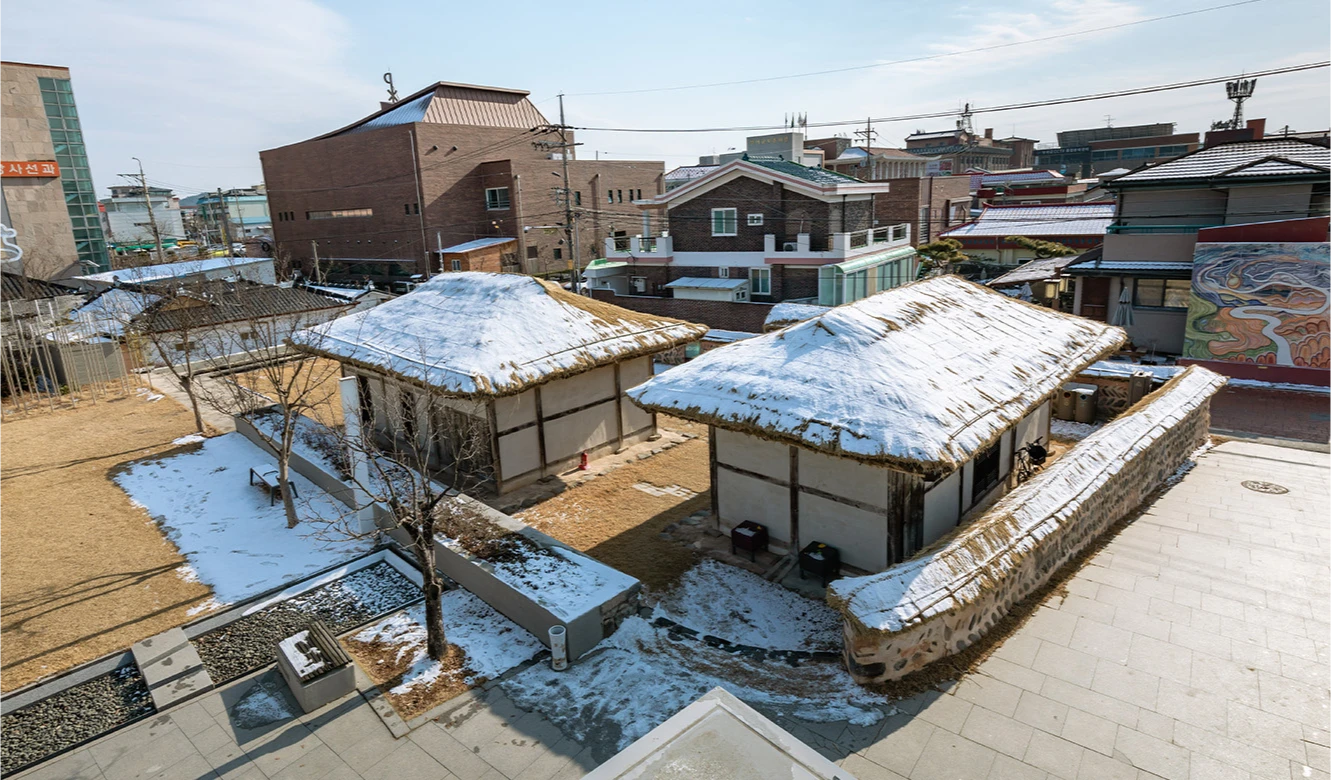 주변 현대식 건물에 둘러싸여 눈이 소복이 쌓인 초가집 두 채가 내려다보이는 신동엽 생가 전경