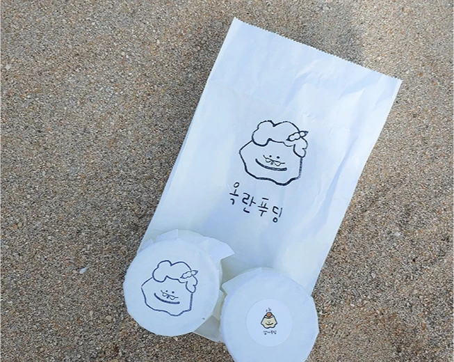 모래 위에 캐릭터가 그려진 종이봉투와 동그란 옥란푸딩 두 개를 올려둔 테이크아웃 모습