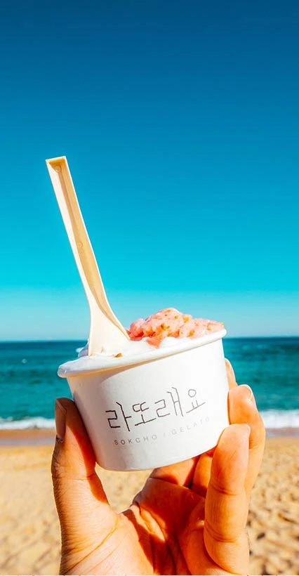 푸른 바다와 모래사장을 배경으로 딸기 토핑을 올린 라또래요 젤라또 컵을 손에 든 모습