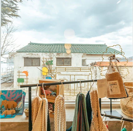 창가에 걸린 손뜨개 소품과 동물 그림 너머로 기와지붕 건물이 보이는 아늑한 매장 진열대