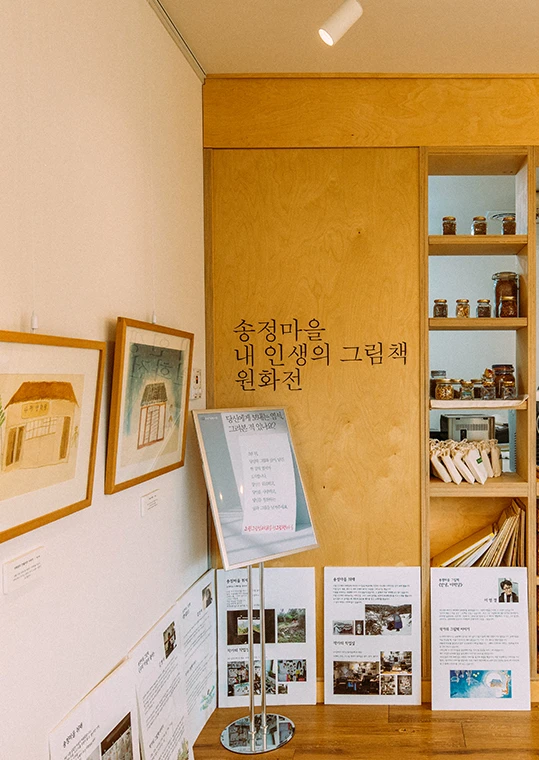 송정마을 내 인생의 그림책 원화전 안내판과 액자 그림이 놓인 전시 공간, 선반에는 병들이 진열돼 있다