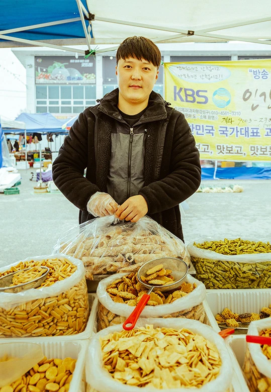 검은 점퍼를 입은 남성 상인이 과자 봉지를 손에 들고 좌판 뒤에 서 있다. 뒤로 KBS 방송 출연 현수막이 걸려 있고 앞에는 각종 과자가 담겨 있다