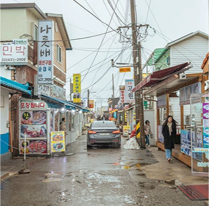 민박과 생선구이 간판이 늘어선 아바이마을 골목, 비 온 뒤 길을 걷는 행인들