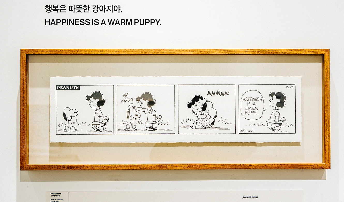 행복은 따뜻한 강아지야 문구 아래 피너츠 네 컷 만화 원화가 액자에 담겨 벽에 걸린 전시