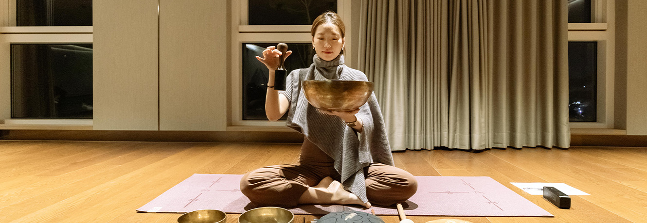 실내에서 회색 숄을 두른 여성이 매트에 앉아 큰 싱잉볼을 들고 말렛으로 소리를 울리고 있다