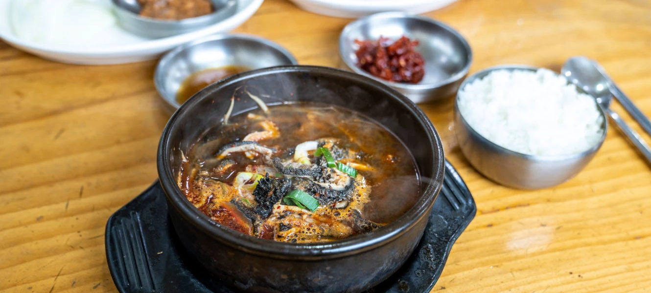 백제해장국집 뚝배기에 담긴 얼큰한 민물생선 해장국과 공기밥, 다진 양념과 밑반찬이 곁들여진 나무 식탁