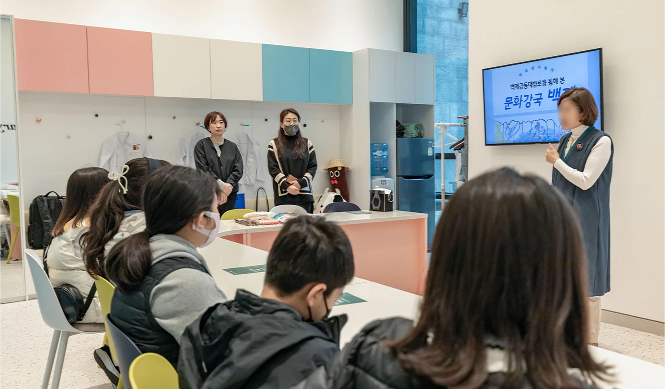 강사가 화면을 가리키며 문화강국 백제를 소개하고 참가자들이 둘러앉아 강의를 듣는 모습