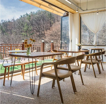 통창 너머로 산과 차밭이 보이는 티하우스 실내, 해바라기 꽃병을 올린 원목 테이블과 의자가 놓여 있다