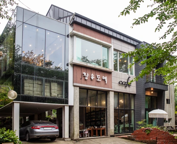 유리와 벽돌로 지은 장유도예 카페 건물 외관, 앞마당에 은색 차와 파라솔이 보이는 모습