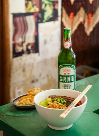 초록빛 테이블 위에 대만식 고기덮밥 한 그릇과 젓가락, 대만 맥주 병, 곁들임 만두 접시가 차려져 있다