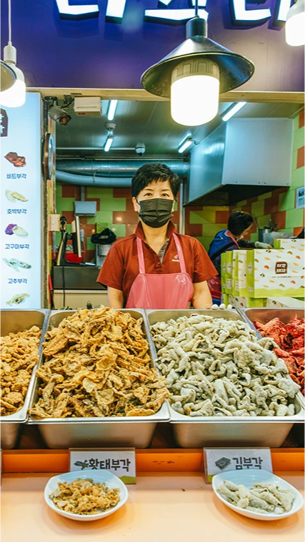 여러 종류의 부각을 진열한 매대 앞에서 앞치마를 두른 상인이 서 있는 티각태각 가게