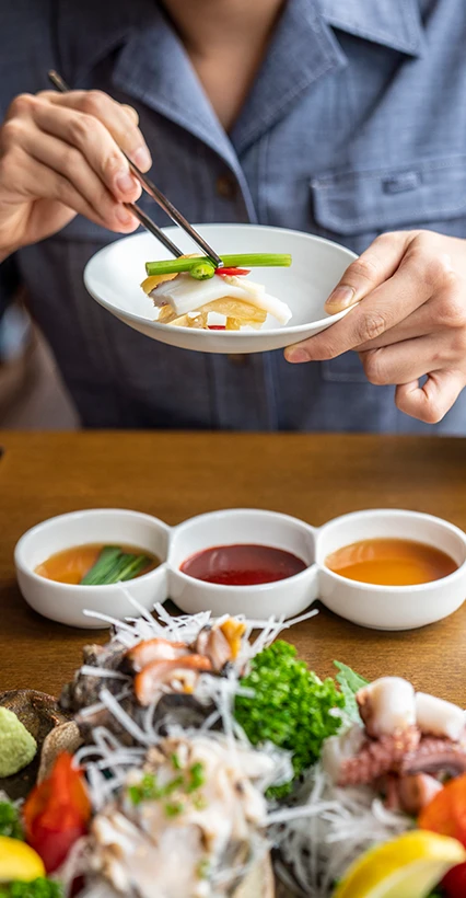 젓가락으로 숙회 한 점을 작은 접시에 덜어 소스를 곁들이는 손, 아래로 모듬 해산물 플래터가 보인다