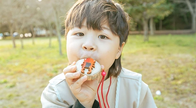 단발머리 아이가 곤충을 색칠한 나무 피리를 입에 물고 정면을 바라보는 모습