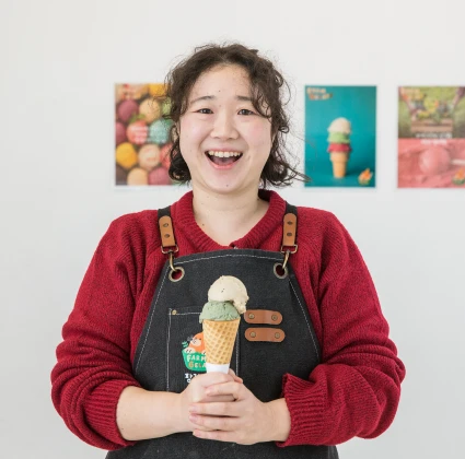 흰 벽 앞에서 젤라또 콘을 들고 활짝 웃는 대표, 뒤로 젤라또 포스터가 걸린 모습