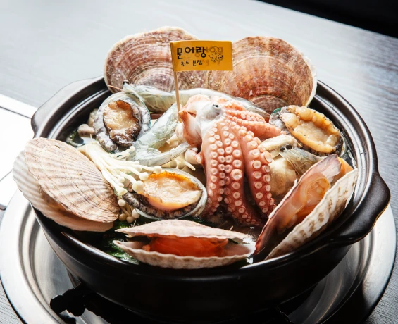 검은 뚝배기에 가리비와 전복, 새우, 문어를 푸짐하게 올린 해신탕에 문어랑 깃발을 꽂은 모습