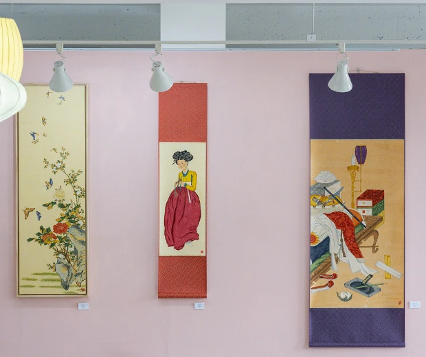 분홍빛 벽에 나란히 걸린 꽃과 여인, 기물을 그린 제주 민화 족자 세 점이 전시된 모습