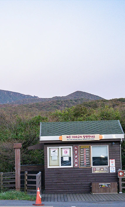 해 질 무렵 부드러운 하늘빛 아래 초록 지붕의 제주 1100고지 탐방안내소 건물과 뒤편으로 완만한 오름이 보이는 풍경