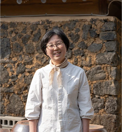 현무암 돌담 앞에서 흰 셔츠에 스카프를 두르고 미소 짓는 고소리 술 익는 집 대표