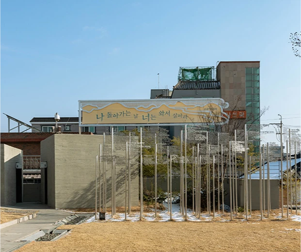 노출 콘크리트 외벽에 시 구절 현수막이 걸리고 마당에 금속 봉 조형물이 선 신동엽문학관 외관